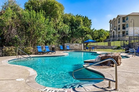 A resort-style pool here at Canyon Vistas with a curved swimming pool, stone water feature, blue lounge chairs, shaded seating, and surrounding greenery in a relaxing outdoor setting.