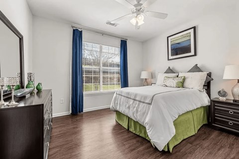 A bright bedroom here at Canyon Vistas with wood-style flooring, a white bed with green accent bedding, bedside tables with lamps, a large window with blue curtains, and a ceiling fan.