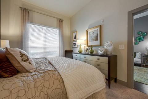 A bedroom here at Cityplace Heights with a neatly made bed in neutral bedding, a dark dresser with green and white decor, framed wall art, a table lamp, and a window with blinds and sheer curtains.