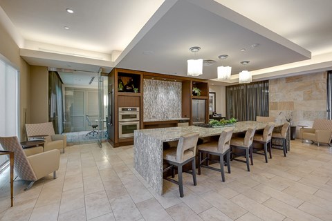 A resident lounge here at Cityplace Heights with a long stone island table, cushioned bar chairs, pendant lights, built-in appliances, tan tile flooring, and nearby seating areas with beige lounge chairs and glass-walled meeting room.