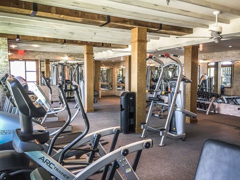 A spacious fitness center here at Crosby at the Brickyard with modern cardio and strength equipment, warm wood beam ceilings, mirrored walls, and bright lighting, creating an inviting and energizing workout space.