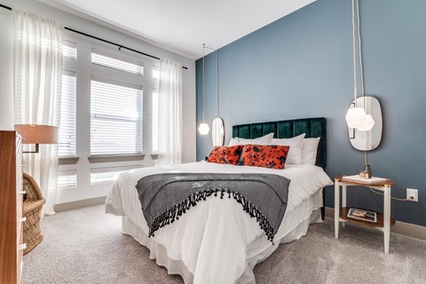 A bright bedroom here at Crosby at the Brickyard with a blue accent wall, plush white bed, bold orange pillows, and large windows, creating a cozy, modern, inviting space.
