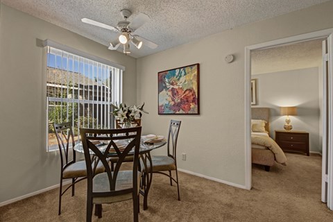A bright dining area here at Cypress Pointe with beige carpet, neutral walls, and a large window with vertical blinds bringing in natural light. A ceiling fan adds comfort, while the open layout offers a clear view into the bedroom, creating a warm and inviting space for everyday dining.