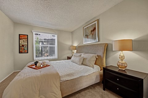 A comfortable bedroom here at Cypress Pointe with plush beige carpet, neutral walls, and a large window with vertical blinds allowing natural light to fill the room. A neatly staged bed with soft bedding and matching bedside lamps creates a calm, cozy, and inviting atmosphere.