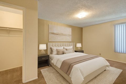 A comfortable bedroom here at Fairgreen with plush beige carpet, soft neutral walls, and a large window with vertical blinds bringing in natural light. A neatly staged bed with neutral bedding and an adjacent walk-in closet create a calm, inviting, and functional retreat.