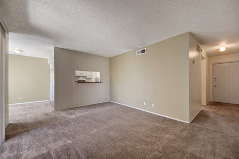 A spacious, empty living room here at Fairgreen with plush beige carpet, soft neutral walls, and an open layout, featuring a pass-through window into the kitchen and a nearby entry hallway that adds to the room’s functional, inviting feel.