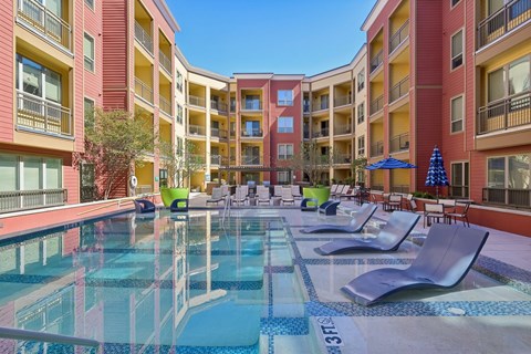 A pool surrounded by chairs and umbrellas in front of apartment buildings.