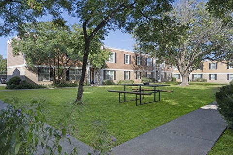 A quiet outdoor courtyard here at Golden Crest with a lush green lawn, shaded picnic table seating, and mature trees, surrounded by two-story apartment buildings and concrete walkways that create a relaxed, community-oriented setting.