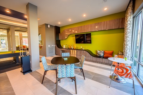 A vibrant community lounge here at Harmony Luxury with modern seating, colorful accent chairs, a built-in refreshment area, and a mounted TV, enhanced by natural light and contemporary design elements that create a fun, social atmosphere.