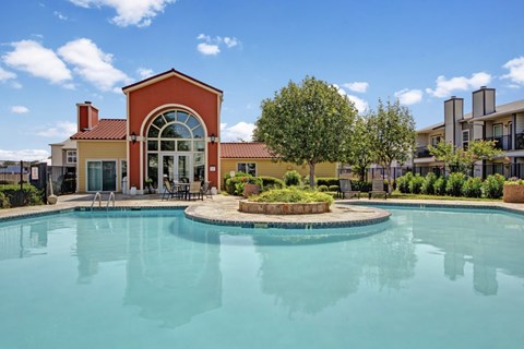 A resort-style swimming pool here at High Plains with clear blue water, surrounding lounge chairs, and a landscaped courtyard, complemented by a clubhouse building and bright skies that create a relaxing, inviting outdoor setting.