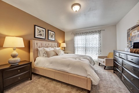 A welcoming bedroom here at High Plains with plush beige carpet, a neutral upholstered bed, warm accent walls, and soft lighting, complemented by a large window with patterned curtains and tasteful furnishings that create a calm, comfortable retreat.