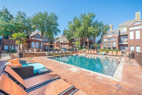 A resort-style outdoor pool here at Lakewood on the Trail with clear blue water, spacious sun deck, lounge chairs, shaded seating areas, and surrounding brick buildings, creating a bright, relaxing community space.