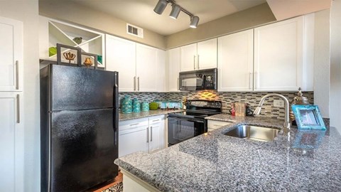 A black refrigerator sits in a kitchen with granite countertops and white cabinets.
