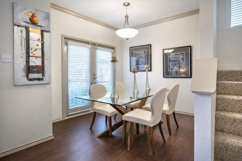 A dining area here at Lakewood on the Trail with a glass-top table, four cream upholstered chairs, silver candle holders, framed wall art, dark wood floors, and bright patio doors with white blinds.