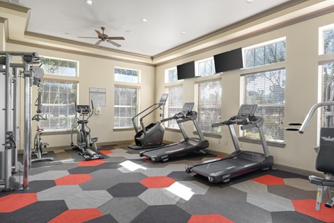 A fitness room here at Landing at Mansfield with treadmills, ellipticals, weight machines, and gray-and-red hex tile flooring, plus large bright windows and wall-mounted TVs.