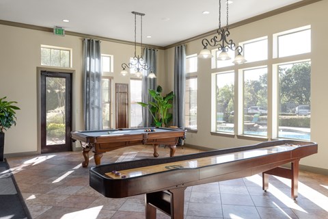 A game room here at Landing at Mansfield with a wooden pool table and shuffleboard table, tall bright windows, dark metal chandeliers, and warm tile flooring overlooking the pool area.