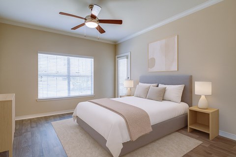 A bedroom here at Landing at Mansfield with a gray upholstered bed, white bedding with beige throw, light wood nightstands, matching white lamps, neutral wall art, and warm wood flooring with a soft cream rug.