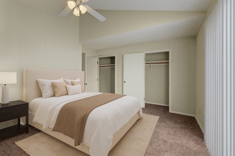 A spacious bedroom here at Las Colinas with soft beige carpet, neutral walls, and a vaulted ceiling with a ceiling fan. Large closets with sliding doors and a bright window with vertical blinds create an airy, comfortable, and inviting space.