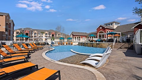 A resort-style outdoor pool here at Latitude 31 featuring a curved swimming pool with in-water lounge chairs, surrounding sun deck lined with orange chaise loungers, shaded seating areas with umbrellas, cascading water features, and upscale apartment buildings creating a vibrant and relaxing community atmosphere.