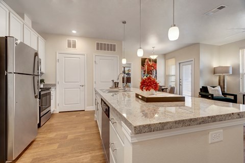 A bright modern kitchen here at Latitude 31 Apartments featuring white cabinetry, stainless steel appliances, a spacious island with a stone-look countertop, pendant lighting, and wood-style flooring that opens into the living area, creating a clean, functional space ideal for cooking, entertaining, and everyday living.