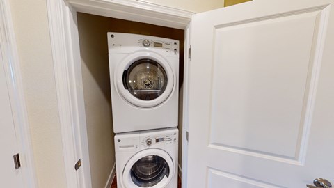 A convenient laundry closet here at Latitude 31 Apartments featuring a stacked front-loading washer and dryer neatly tucked behind a white paneled door. Neutral walls, clean trim, and a compact layout provide an efficient in-home laundry solution that maximizes space while keeping appliances discreet and easily accessible.