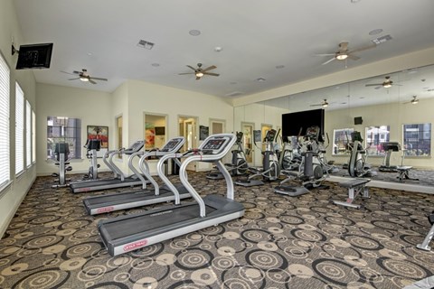 A spacious fitness center here at Le Mirage featuring rows of modern treadmills and cardio machines, mirrored walls, wall-mounted televisions, ceiling fans, patterned flooring, and large windows that bring in natural light for a bright and motivating workout environment.