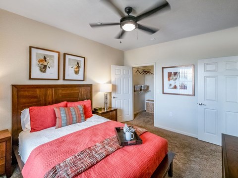 A cozy primary bedroom here at Le Mirage featuring a wooden bed frame with coral bedding and layered pillows, soft carpet flooring, a ceiling fan with light, matching bedside tables with lamps, framed wall art, and an open walk-in closet with shelving and hanging space visible through a white door.