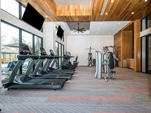 A modern fitness center here at Magnolia on the Green with treadmills, strength machines, high ceilings with wood accents, wall-mounted TVs, and large windows bringing in natural light for an open, energizing workout space.