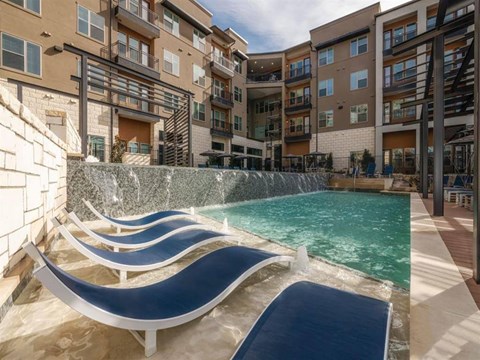 A resort-style pool area here at Magnolia on the Green with in-water lounge chairs, a cascading water feature wall, surrounding apartment buildings with balconies, and sunny deck space for relaxing outdoor enjoyment.
