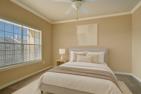 A cozy and serene bedroom here at Mission Green featuring a neatly made bed with white bedding, layered neutral pillows, and a soft throw blanket, complemented by warm beige walls, plush carpeting, a ceiling fan with light, simple abstract wall art above the headboard, and natural light filtering through wide window blinds.