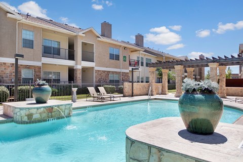 A resort-style outdoor pool area here at Mission Green showcasing clear blue water, a spacious stone pool deck, large decorative planters, shaded pergola columns, lounge chairs, and surrounding apartment buildings under a bright sky, creating a relaxing community space for residents to unwind.