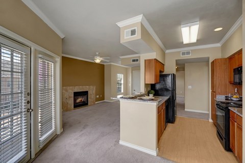 A bright and open living area here at Mission Green showcasing an open-concept layout with a connected kitchen and living space, warm neutral walls, wood-style flooring in the kitchen, soft carpeted living area, a tiled fireplace, ceiling fan, and large windows and glass doors that allow natural light to fill the room.