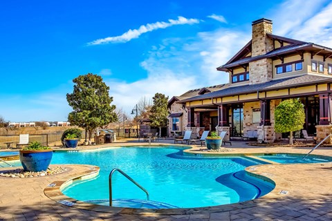 An outdoor pool area here at Monterra Village with a curved blue pool, tan stone deck, large blue planters, lounge chairs, and a stone clubhouse with covered patio seating under a bright sky.