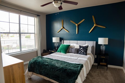 A cozy bedroom here at Monterra Village with soft carpet, a large bed with green and neutral bedding, a bold blue accent wall with decorative art, bedside lamps, and a bright window with blinds.