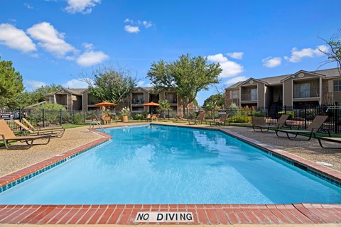 A sunny outdoor swimming pool here at Northridge Court surrounded by a spacious pool deck with lounge chairs, patio umbrellas, and landscaped greenery, with nearby apartment buildings, mature trees, and a clear blue sky creating a relaxing community setting.