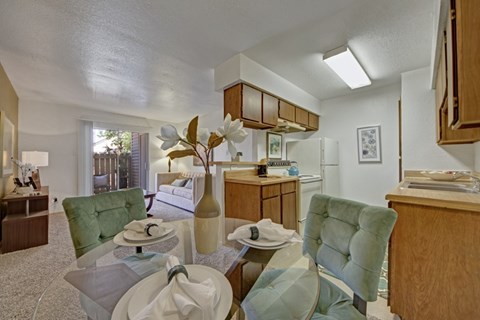 A bright dining and kitchen space here at Northridge Court showcasing warm wood cabinetry, light countertops, and a glass dining table with upholstered chairs, opening into a comfortable living area with soft carpeting, neutral walls, and a sliding glass door that brings in natural light.