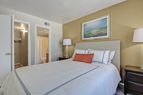 A cozy bedroom here at Northridge Court featuring a queen-size bed with crisp white bedding and accent pillows, a soft upholstered headboard, matching nightstands with lamps, warm neutral walls, framed artwork above the bed, carpeted flooring, and an open doorway leading to a private bathroom and closet area.