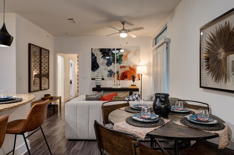 A dining and living area here at Reserve at Arboretum with a round dark wood table, brown leather chairs, a beige sofa with colorful accent pillows, and warm modern artwork, creating a bright, stylish, and welcoming open space.