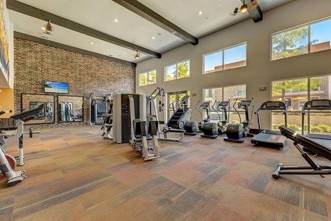 A fitness center here at Skyline Farmers Market with large windows, brick accent wall, treadmills and ellipticals, strength machines, mirrors, and high ceilings with exposed beams and bright natural light.
