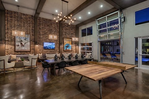 A resident lounge here at Skyline Farmers Market with brick accent walls, dark wood beams, long community table, wood table tennis table, warm chandelier lighting, and modern seating areas opening to an outdoor pool patio.