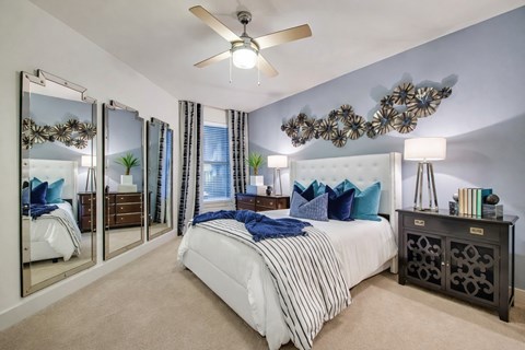 A bedroom here at Skyline Farmers Market with a white tufted bed, blue and teal accent pillows, navy throw blanket, mirrored wall panels, black nightstands, and soft neutral carpet under bright lighting.