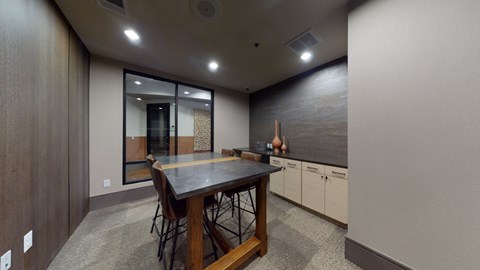 A work lounge here at Sorrel Fairview with a tall dark wood table, brown leather stools, stone-look tabletop, light wood cabinets, and a sleek accent wall, creating a modern, quiet meeting space.