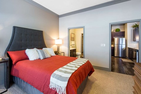 A bedroom here at Sorrel Fairview with a dark gray tufted headboard, red bedspread, white accent pillows, striped throw blanket, soft carpet flooring, and warm bedside lamp lighting.