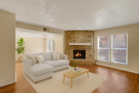 A bright, inviting living room here at Summertree Place featuring warm wood-style flooring, a neutral sectional sofa with soft pillows, a light area rug, and a simple coffee table, complemented by a brick fireplace, large windows with blinds, and an open layout that allows natural light to fill the space.