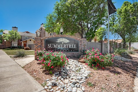A welcoming monument sign here at Summertree Place showcasing a brick-framed community sign surrounded by blooming pink flowers, decorative stone landscaping, mature trees, and nearby apartment buildings, creating a polished and inviting entrance along a sunny residential sidewalk.