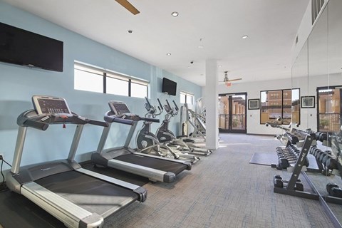 A fitness center here at The Uptown with modern cardio machines, treadmills and ellipticals, a mirrored wall with dumbbell rack, blue accent wall, and bright natural light from tall windows.