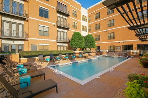 A pool courtyard here at The Uptown with a rectangular swimming pool, brick deck, black lounge chairs, bright teal side tables, trimmed green trees, and surrounding mid-rise brick apartment buildings in sunny light.