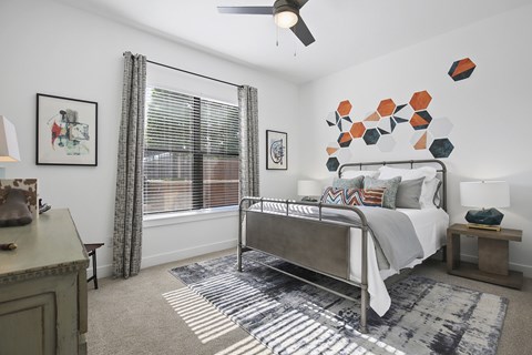 A bedroom here at The Uptown with a gray metal bed frame, white bedding, colorful orange and teal accent pillows, geometric wall art, patterned gray rug, wood nightstand, and bright window with striped curtains.