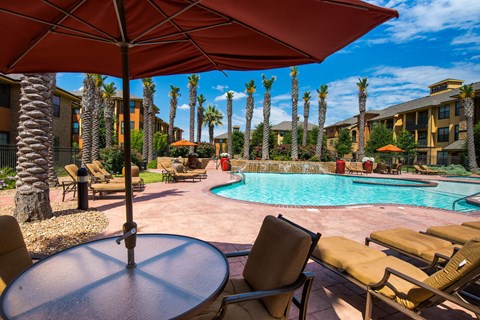 A resort-style outdoor pool area here at Tuscany at Faudree featuring a sparkling curved swimming pool surrounded by palm trees, tan lounge chairs, and shaded seating under large umbrellas. Warm-toned buildings, landscaped greenery, and a sunny blue sky create a relaxing, vacation-inspired community atmosphere.