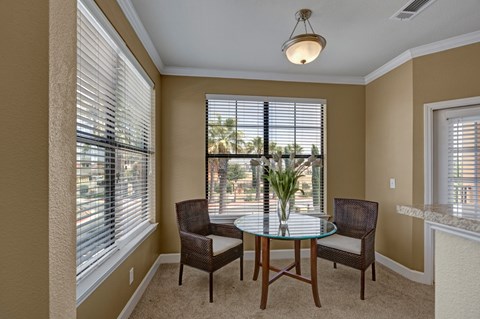 A bright dining nook here at Tuscany at Faudree with plush carpet and neutral walls. A small round glass-top table with two chairs sits beneath a ceiling light, while large windows with blinds bring in natural light and views of surrounding greenery, creating a cozy space for everyday meals.